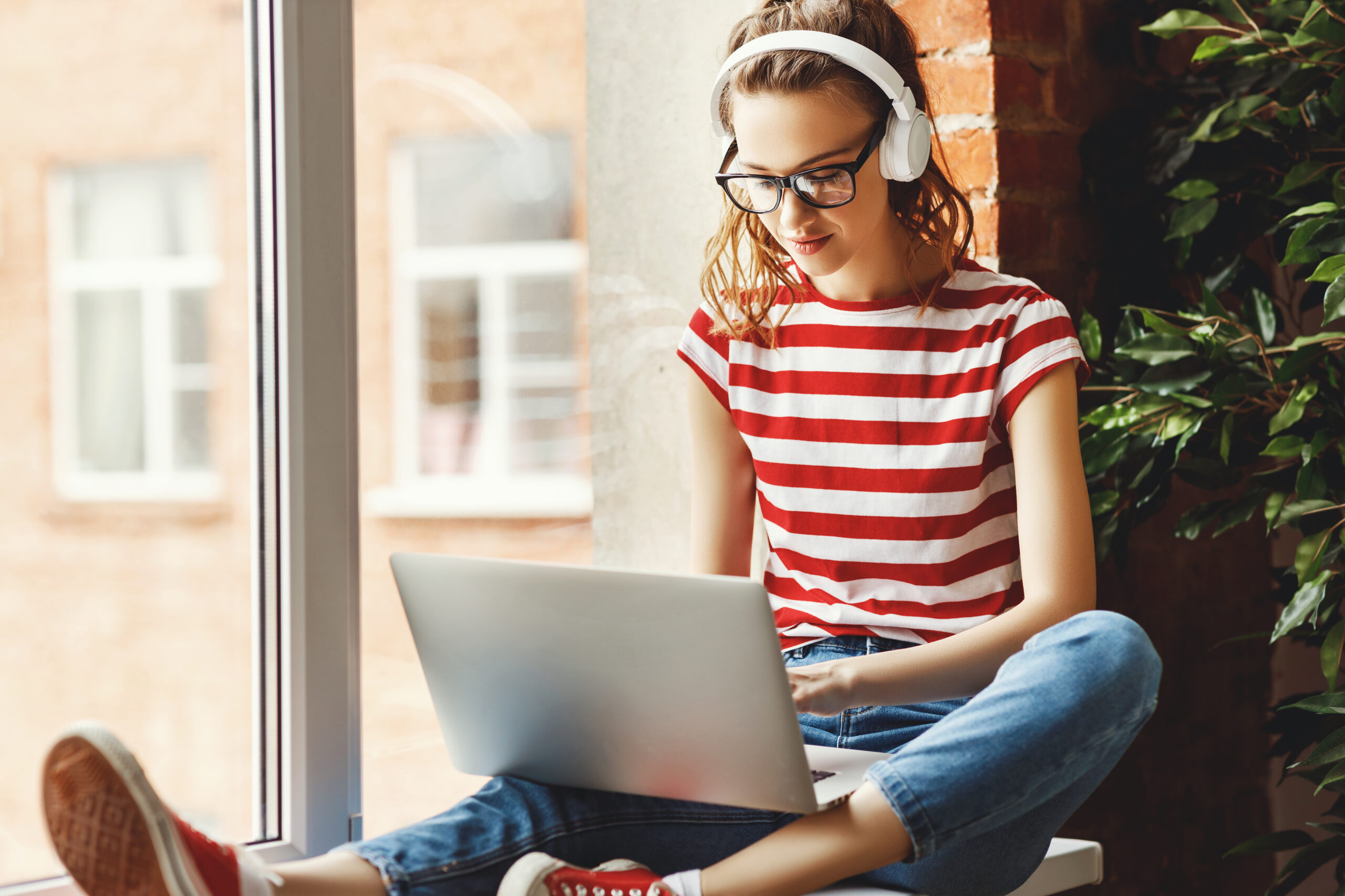 Crop pensive delighted female in glasses and wireless headphones focusing on screen and interacting with laptop while sitting on window sill near green potted tree in loft office