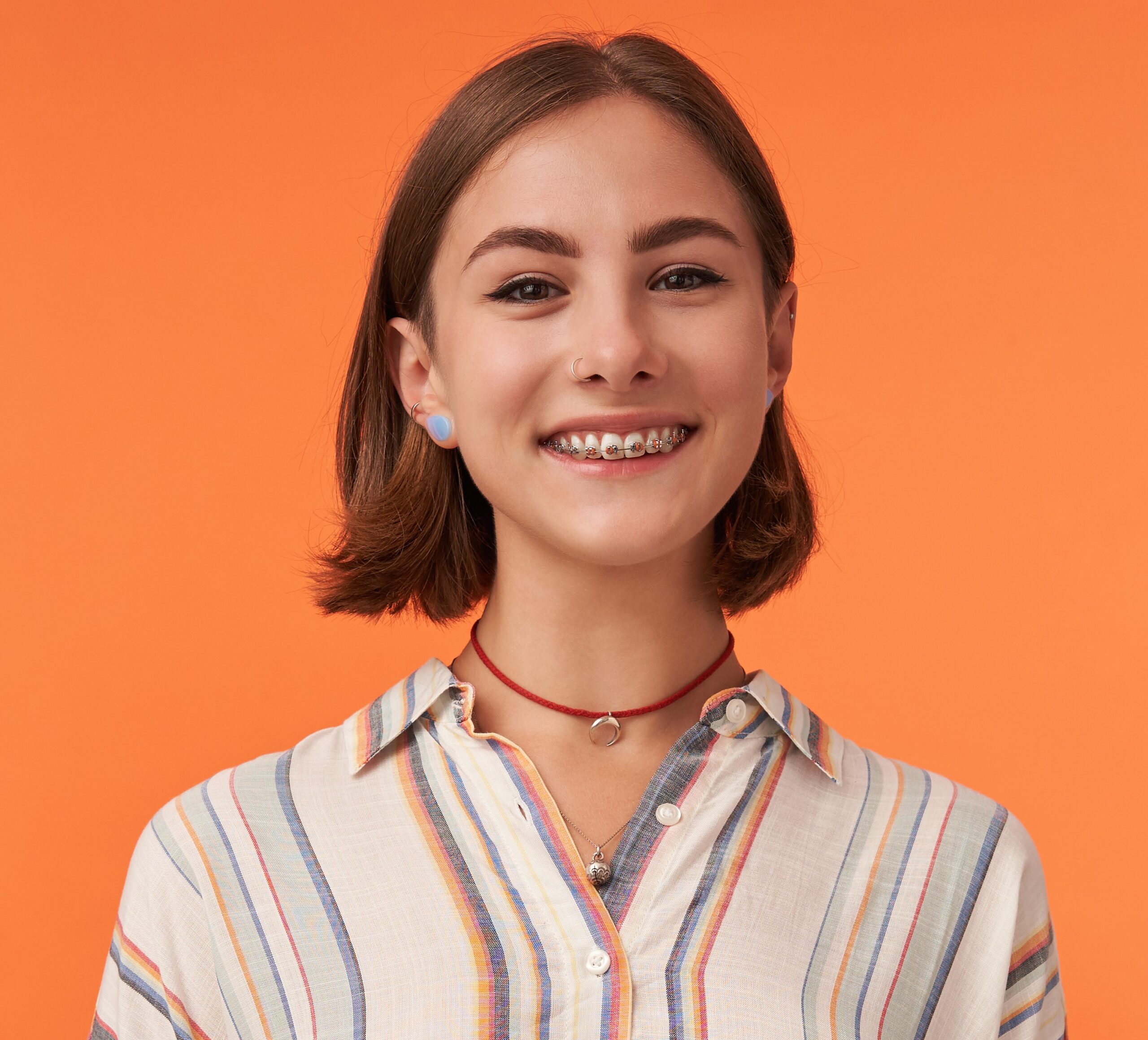 Portrait of girl with brown short haircut and braces for teeth, pierced nose, wearing striped shirt. Young smiling girl watching camera against orange background