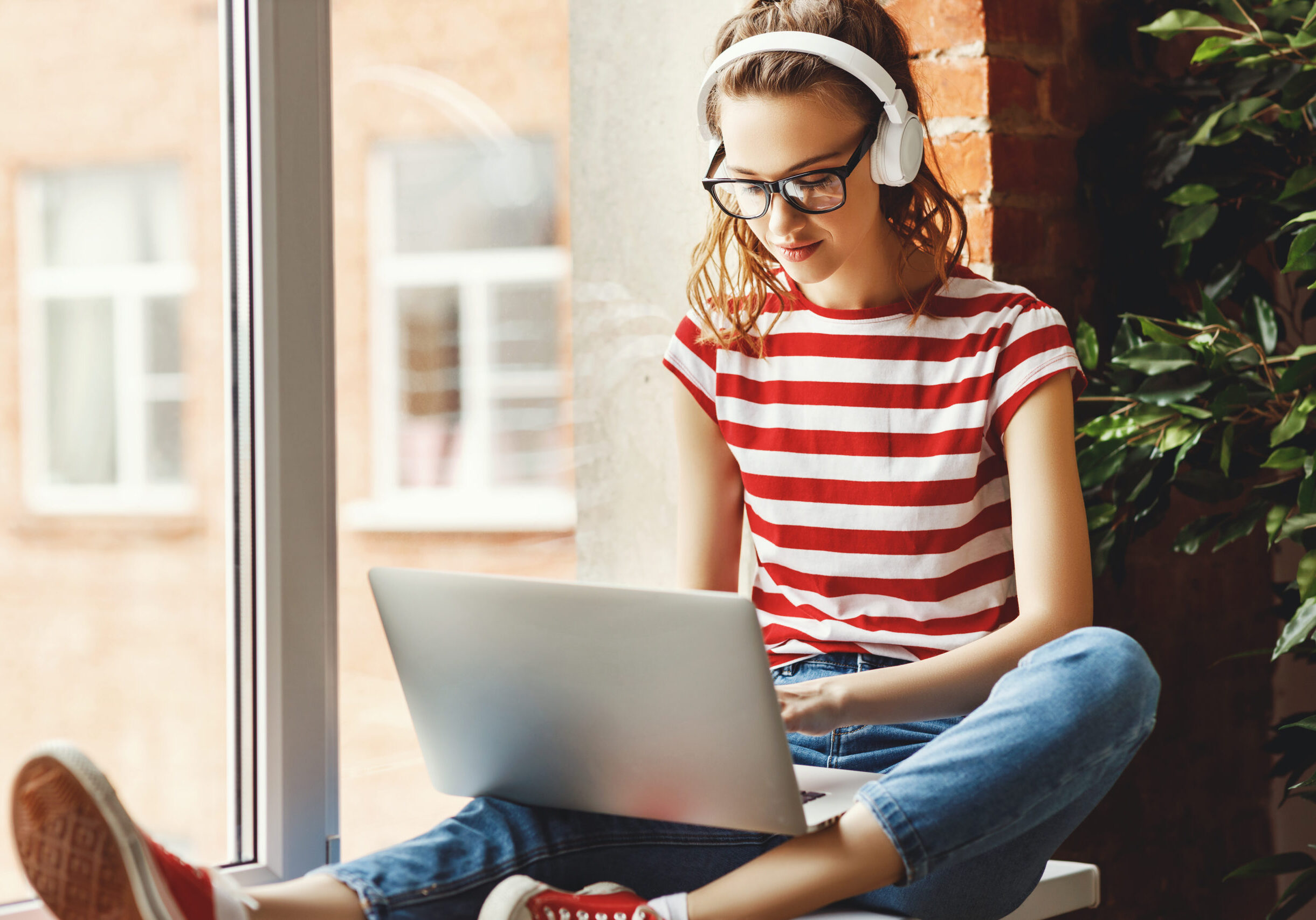 Crop pensive delighted female in glasses and wireless headphones focusing on screen and interacting with laptop while sitting on window sill near green potted tree in loft office