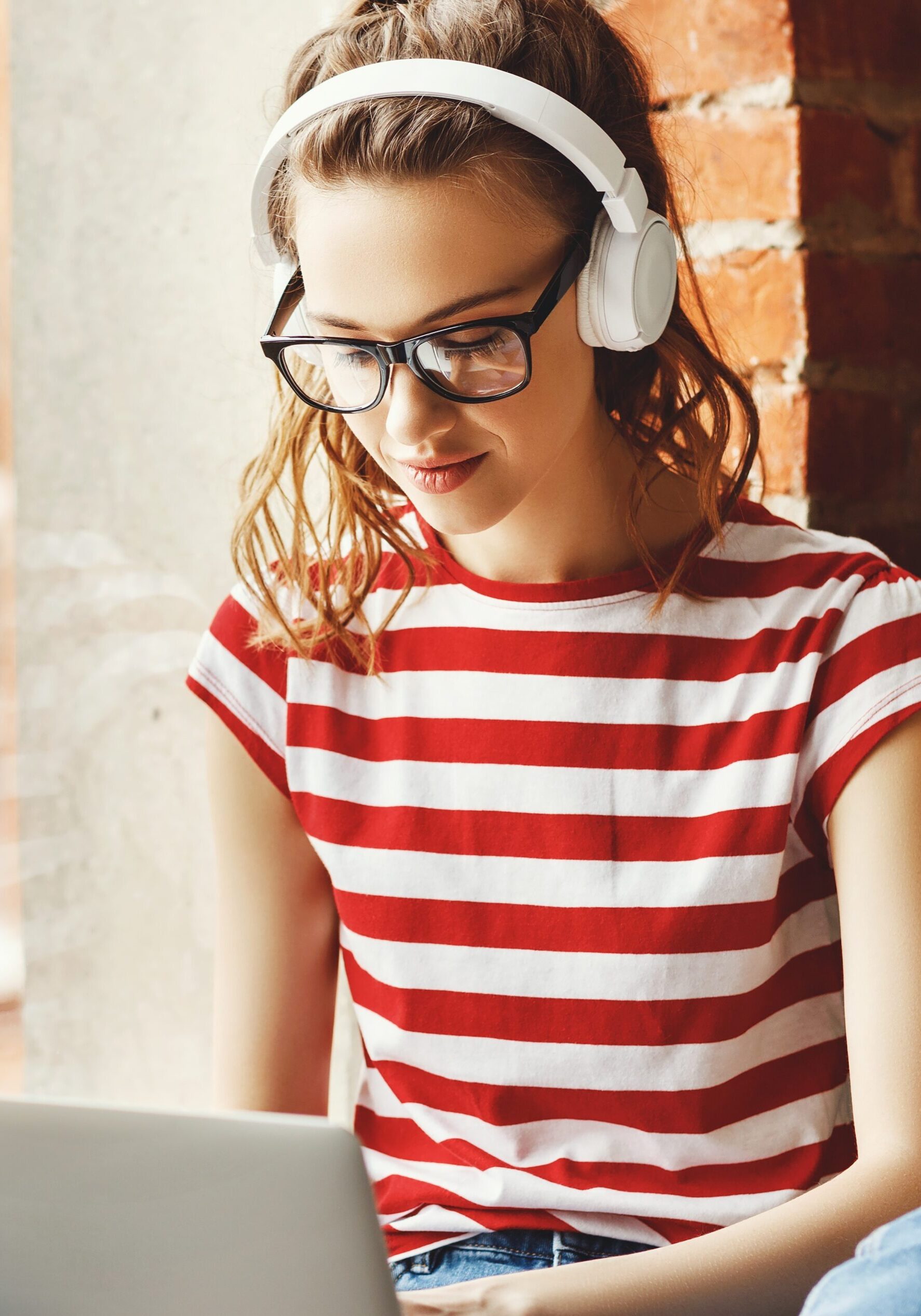 Crop pensive delighted female in glasses and wireless headphones focusing on screen and interacting with laptop while sitting on window sill near green potted tree in loft office