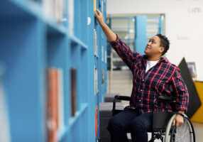 Portrait of disabled student in wheelchair choosing books while studying in college library, copy space