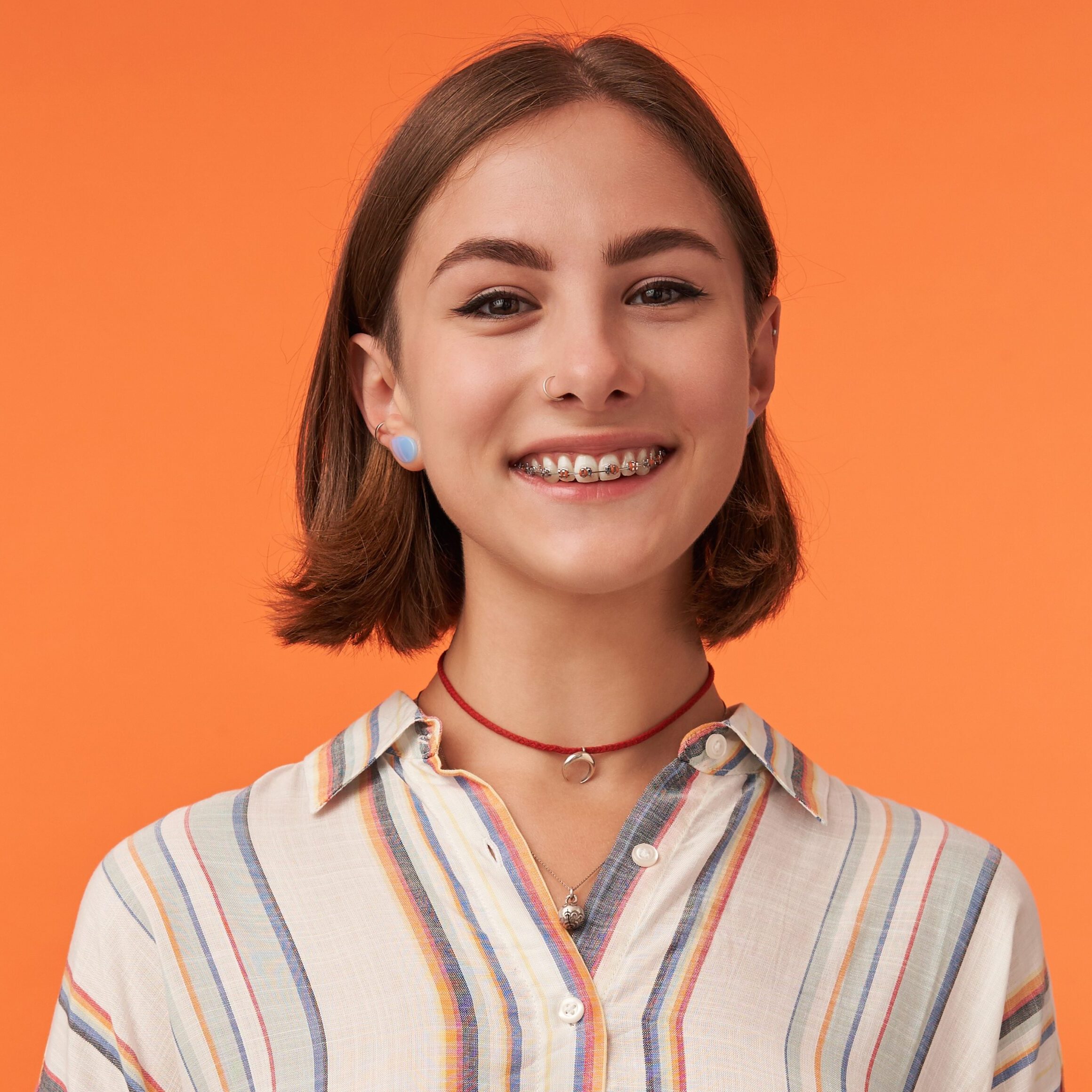 Teen girl 3 crop Portrait of girl with brown short haircut and braces for teeth, pierced nose, wearing striped shirt. Young smiling girl watching camera against orange background